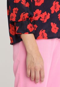 Blusa blu navy con motivi floreali rossi, con polsini arricciati. Abbinata a pantaloni rosa chiaro, che mostrano una texture di tessuto liscia.