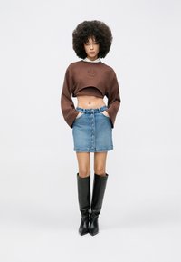 Braunes, kurz geschnittenes Sweatshirt mit geprägtem Detail, kombiniert mit einem hellblauen Denimrock mit Knopfleiste vorne. Dazu schwarze kniehohe Stiefel getragen.