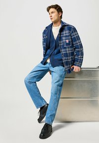 Veste à carreaux marine avec des boutons, superposée à un gilet texturé bleu et une chemise crème, associée à un jean bleu clair et des chaussures noires.