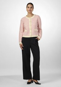 Cardigan tricoté en rose clair avec un motif à carreaux, accents crème, encolure ronde, fermeture à boutons et deux poches avant. Assorti à un pantalon noir.
