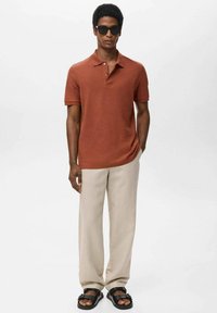 Ein rostfarbendes Poloshirt mit kurzen Ärmeln, kombiniert mit hellbeigen Hosen. Das Modell trägt schwarze Sonnenbrillen und Sandalen mit Riemen.