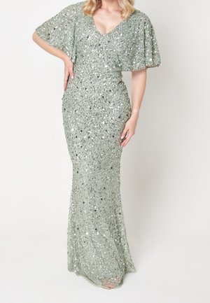 Kvinde iført en gulvlange sølvglitrende kjole med flagrende ærmer og V-udskæring, stående med den ene hånd på hoften.