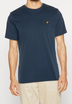 Mężczyzna ubrany w granatowy t-shirt z krótkim rękawem z małym żółtym logo ptaka na lewej piersi oraz beżowe spodnie.