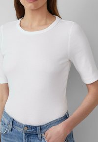 Top corto a maniche corte bianco con scollatura rotonda, abbinato a jeans in denim azzurro chiaro con chiusura a bottone e tasche visibili.