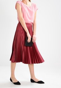 Hellrosa Kurzarmoberteil kombiniert mit einem tiefroten, plissierten Midirock. Eine schwarze Clutch und spitze schwarze Ballerinas runden das Outfit ab.