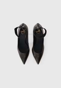 Pantofi cu toc înalt din piele neagră, cu vârf ascuțit, închidere cu fermoar și o brățară cu un logo de inimă aurie la gleznă.