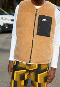 En tanfärgad fleeceväst med svart dragkedjefack och logotyp, kombinerad med mönstrade gula och gröna byxor som framhäver geometriska former.