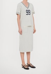 Proužkovaný dvoudílný komplet zahrnující krémový top s námořnicky modrými pruhy a číslem "99", doplněný o odpovídající pouzdrovou sukni a černé baleríny.