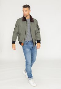Veste bomber vert olive avec col en fausse fourrure marron, poignets côtelés et une étiquette orange sur le côté gauche. Portée avec un jean bleu clair et des baskets blanches.