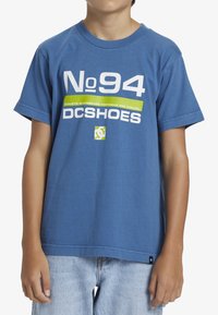 Modrá bavlněná tričko s výrazným bílým a zeleným textem: "Č. 94 ATLETICKÁ SKATEBOARDOVÁ OBUV A OBLEČENÍ DC SHOES." Krátké rukávy.