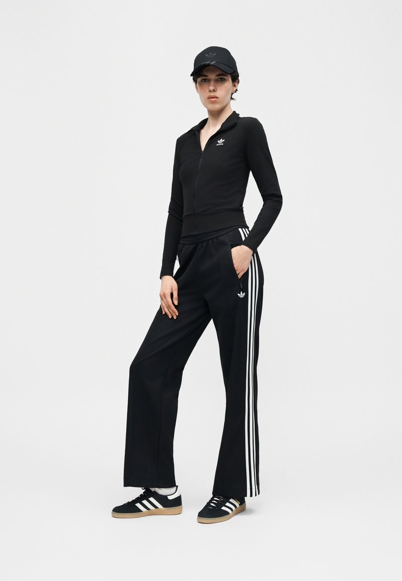 Femme debout, une main dans la poche, portant un survêtement Adidas noir, une casquette et des baskets avec des bandes blanches, sur un fond uni.