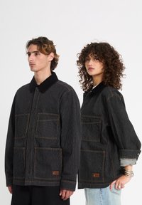 Deux personnes portant des vestes noires à fines rayures avec de grandes poches avant, debout côte à côte sur un fond blanc uni.