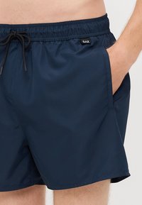 Námorníckomodré plavky vyrobené z hladkej, ľahkej tkaniny. Obsahujú elastický pás so šnúrkou a bočné vrecká. Jemný detail loga.