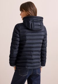 Giacca puffa blu navy con cappuccio, caratterizzata da un design a righe orizzontali, polsini elasticizzati e colletto strutturato. Indossata sopra pantaloni in denim.