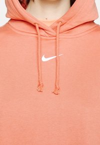 Ljust korallfärgad hoodie med dragsko, med en vit Nike-logga på bröstet och mjukt, texturerat tyg.