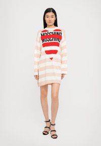 Abito oversize a righe in rosa chiaro e bianco, con una grafiche audaci in rosso e nero con il logo "Moschino" e un design a cuore. Maniche lunghe e vestibilità rilassata.