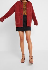 Czerwona kurtka z płótna z przednimi kieszeniami i białymi przeszyciami, noszona na wzorzystej bluzce. Połączona z czarną dżinsową mini spódnicą i sandałami na obcasach.