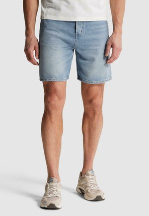 Muž oblečený v svetlomodrých džínsových kraťasoch, bielej tričku a béžových športových topánkach, stojaci pred jednoduchým svetlým pozadím.