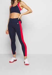 Kvinde iført navy blue sports-bh og leggings med røde og hvide striber, som poserer med den ene hånd på hoften og hvide løbesko.