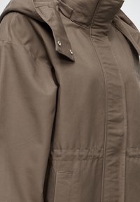Gros plan d'une veste marron avec rabat à bouton-pression, col montant et coutures visibles sur la poche avant.
