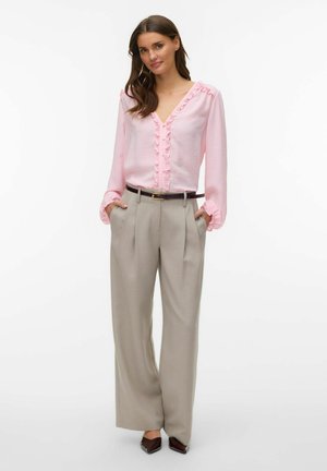 Vrouw met een lichtroze blouse met ruches, gestopt in beige wijde broek met hoge taille, een zwarte riem en bruine puntige schoenen.