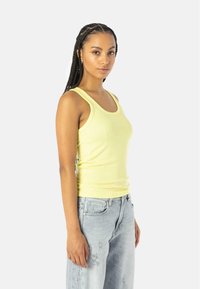 Kollane tank top, mis on valmistatud pehmest ja venivast materjalist, avara kaelusega. Kombineeritud heledate hallide kulunud teksadega. Lihtne disain, ilma mustriteta.