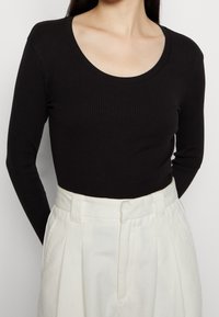 Maglietta nera a maniche lunghe con scollatura a barca, abbinata a pantaloni bianchi a pieghe a vita alta, che mette in risalto il contrasto di texture e design.