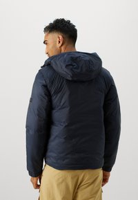 Doudoune marine avec capuche, présentant une surface lisse et brillante ainsi que des poignets élastiques. La vue arrière montre un badge de logo sur la manche gauche.