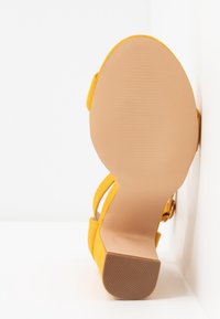 Sandale en daim jaune avec une semelle beige lisse et un petit talon bloc, vue de dessous sur un fond blanc.