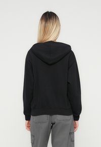Person med langt blondt hår iført en svart hettegenser og grå cargo-bukser, vendt mot en hvit vegg med hendene avslappet ved sidene.