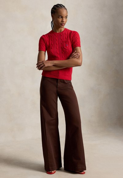 Kobieta z zaplecionymi włosami, ubrana w czerwony sweter z krótkim rękawem ze splotem warkoczowym i brązowe spodnie o szerokich nogawkach, stojąca z założonymi ramionami.