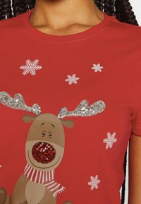 Camiseta roja con un reno de dibujos animados que tiene cuernos de lentejuelas plateadas, una nariz de lentejuelas rojas y una bufanda a rayas, acompañada de copos de nieve blancos.