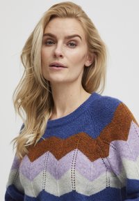 Gebreide trui met een ronde hals, voorzien van een zigzagpatroon in blauw, oranje, lavendel en crème. Zachte textuur met open steekdetails.