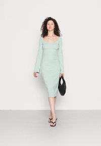 Vestido justo verde menta com decote arredondado e mangas longas; até aos joelhos. Modelo segura uma mala preta acolchoada e usa sandálias pretas.