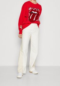 Rotes Sweatshirt mit langen Ärmeln, versehen mit einem großen Rolling-Stones-Zungenlogo, kombiniert mit weißen Hosen mit weitem Bein und weißen Turnschuhen.