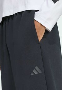 Pantalons de sport gris foncé avec une texture lisse, dotés de poches latérales et d'un accent logo noir subtil sur le bas de la jambe gauche.