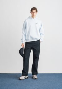 Sweat à capuche bleu clair avec une poche avant, pantalon noir et chaussures de sport grises. Une casquette noire est tenue dans une main.