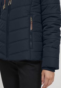 Veste matelassée marine avec une poche zippée, coutures visibles et un col en polaire. Elle présente un design ajusté et des poignets élastiques.