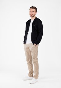 Hombre con las manos en los bolsillos, vistiendo chaqueta negra con cremallera, camiseta blanca, pantalones beige y zapatillas blancas, de pie sobre un fondo blanco.