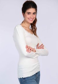 Weißes, geripptes Langarmshirt mit Wellenausschnitt und -ärmeln, aus weichem Material, kombiniert mit blauen Jeans.