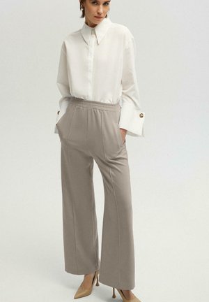 Femme portant une chemise blanche oversize à manches retroussées, un pantalon beige taille haute à jambes larges et des escarpins beiges à bouts pointus, debout les mains dans les poches.
