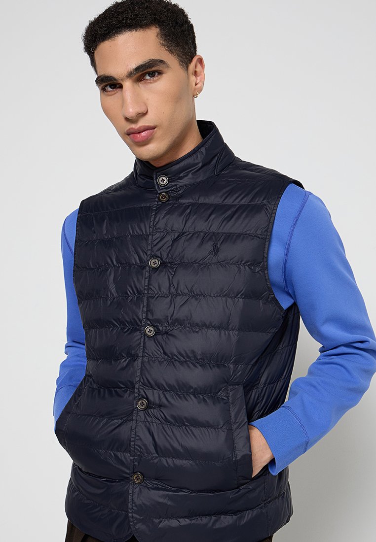 Granatowa kamizelka puchowa z wysokim kołnierzem, zapięciem na guziki z przodu i bocznymi kieszeniami. Nosi się ją na niebieskiej koszuli z długim rękawem. Gładka tekstura.
