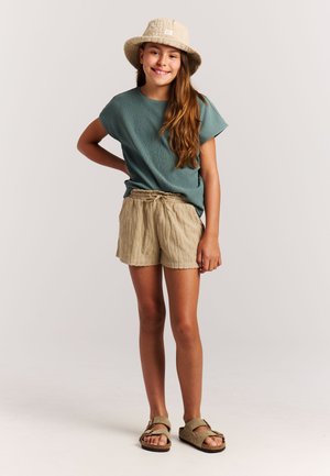 Jeune fille souriante debout, main sur la hanche, portant un chapeau bob beige, un haut à manches courtes bleu sarcelle, un short beige et des sandales beige sur fond blanc.