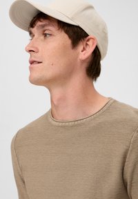 Beige katoenen crew neck shirt met een ribgebreide kraag, gecombineerd met een lichtbruine pet. Weergegeven vanuit een zijprofiel.
