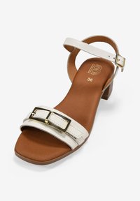 Sandalia de tacón de cuero blanco con dos correas, una de ellas con una hebilla dorada. Plantilla acolchada marrón, forma de punta rectangular y tacón cuadrado.
