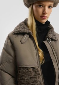 Myoung woman z długimi blond włosami, nosząca brązową puchową kurtkę z kołnierzem z owczej wełny i pasującą futrzaną czapkę na czarnym golfie.