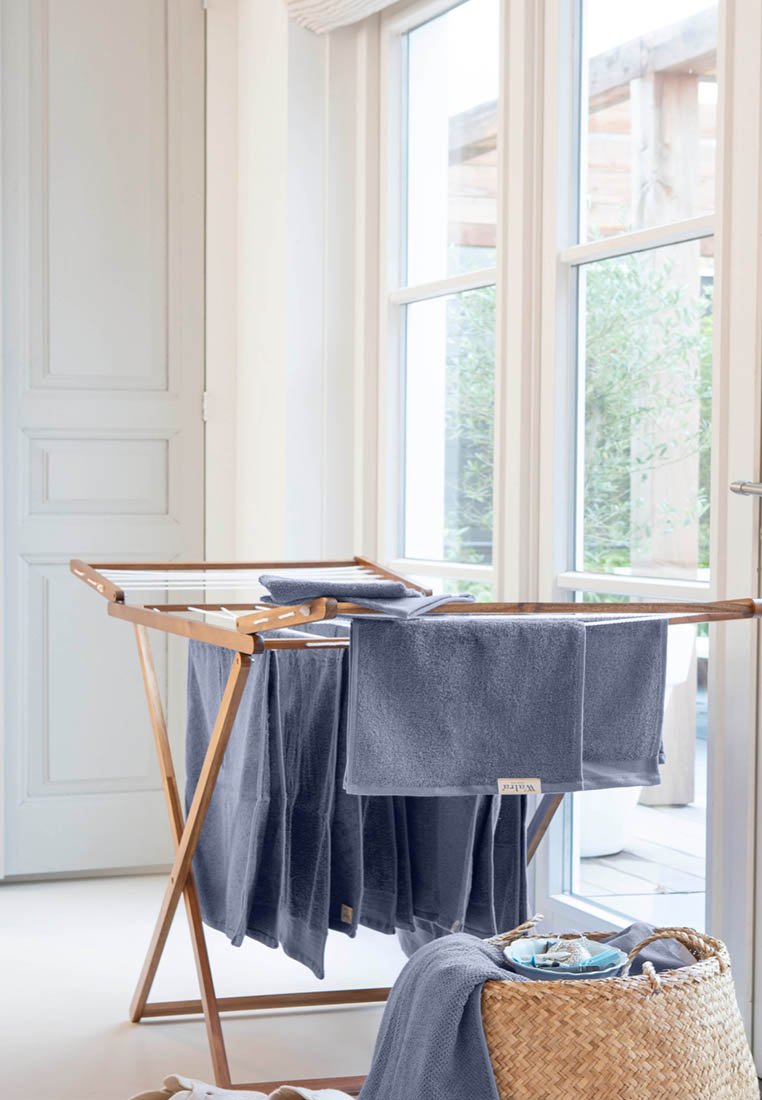 Een houten droogrek houdt verschillende donkergrijze handdoeken vast, met een gestructureerde stof. Daaronder staat een gevlochten mand, met kleinere handdoeken en schalen.