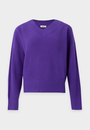 Femme aux longs cheveux foncés portant un pull violet à col en V, un pantalon gris à carreaux taille haute avec de petits pois, une ceinture foncée et des mocassins noirs.