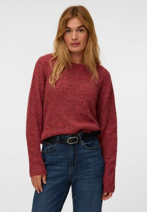 Femme aux longs cheveux blonds portant un pull en maille rouge rentré dans un jean bleu avec une ceinture noire, debout devant un fond clair uni.