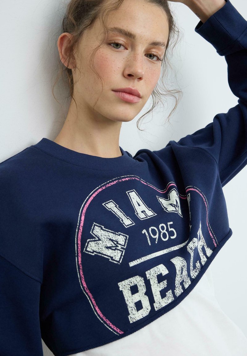 Jeune femme avec de légères taches de rousseur portant un sweat-shirt bleu marine et blanc avec le texte "Miami Beach 1985", levant un bras.